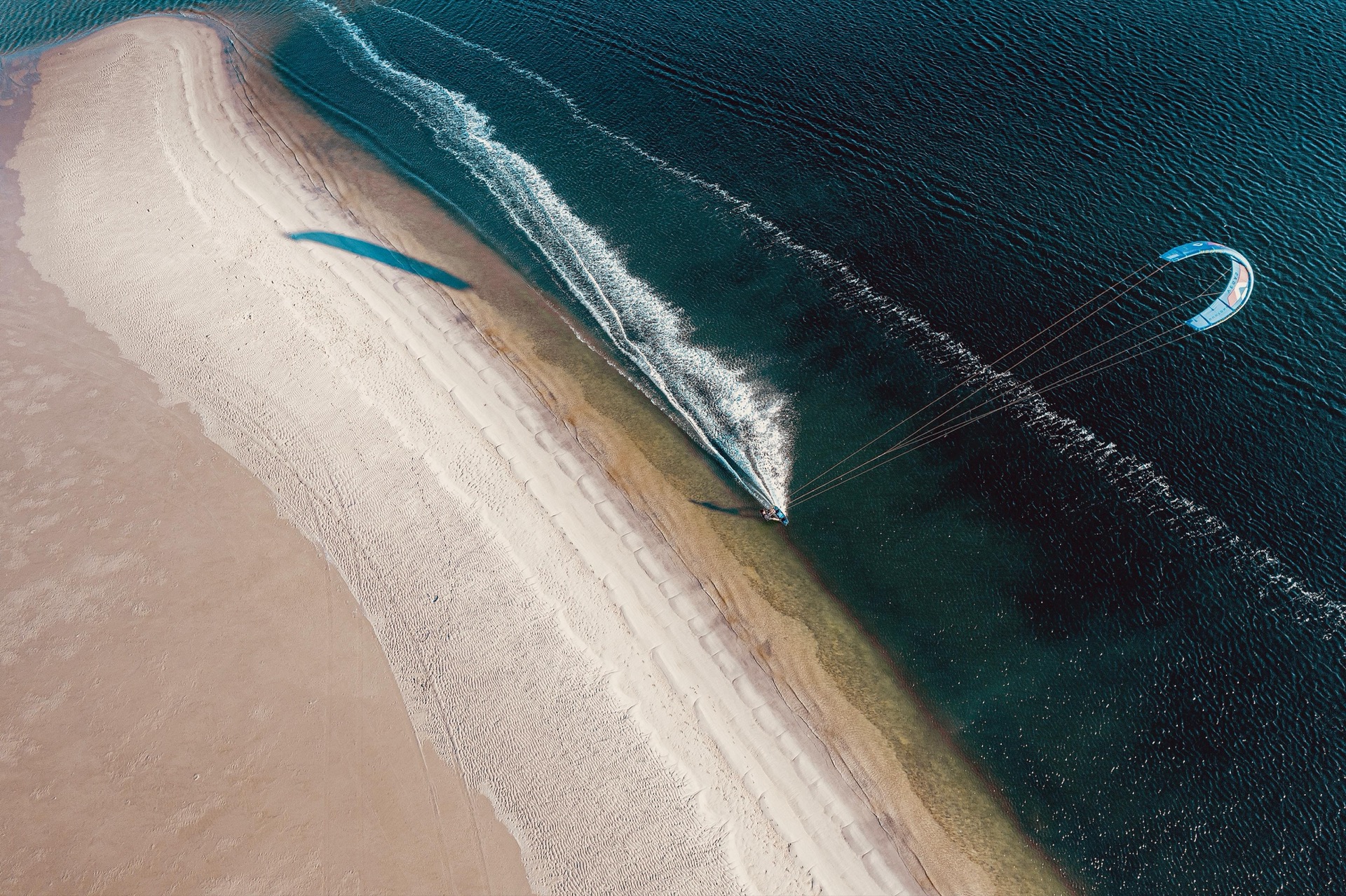 Kitesurfer racing along the sand peninsula leaving a spray trail, aerial view